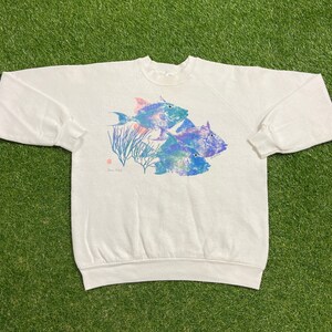 以下が含まれることがあります： 白のスウェットシャツ。青、紫、ターコイズ色の3匹の魚のカラフルなグラフィックが特徴です。デザインにはサンゴと小さな赤いスタンプが含まれています。スウェットシャツは長袖で、クルーネックです。