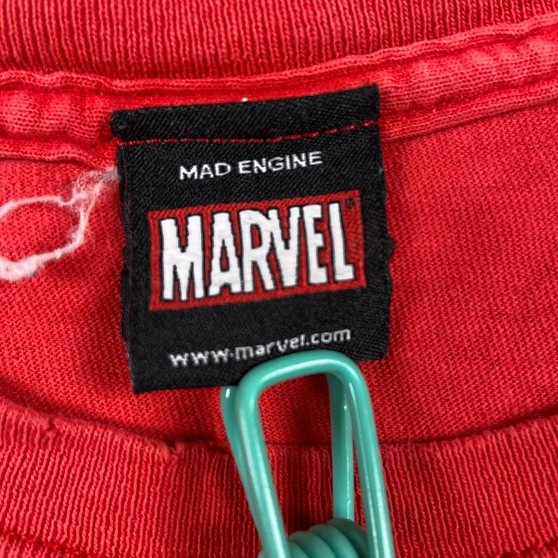Puede incluir: Una etiqueta en blanco y negro con el texto "MAD ENGINE" y "MARVEL" en letras rojas. La etiqueta tambi&eacute;n incluye el sitio web "www.marvel.com".