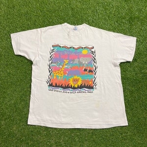 May include: White t-shirt featuring a colorful graphic of African animals, including a giraffe, zebra, and elephant. The text "SAN DIEGO ZOO & WILD ANIMAL PARK" is printed below the graphic. The shirt is on a green surface.