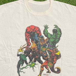 Op de afbeelding: Wit T-shirt met een kleurrijke grafische print van meerdere superhelden die tegen Thanos vechten. De grafiek bevat Hulk, Thing, Green Lantern en anderen. De grafiek is gesigneerd met "Jim Calafiore 97".