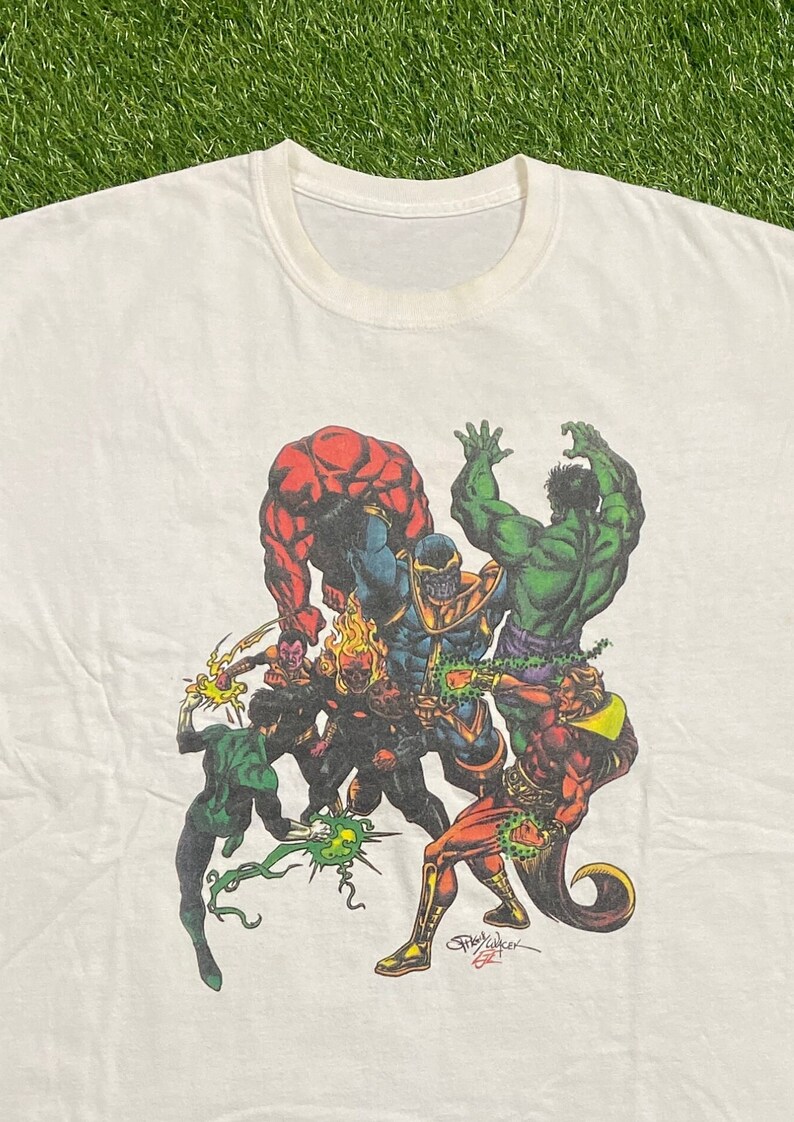 Op de afbeelding: Wit T-shirt met een kleurrijke grafische print van meerdere superhelden die tegen Thanos vechten. De grafiek bevat Hulk, Thing, Green Lantern en anderen. De grafiek is gesigneerd met "Jim Calafiore 97".