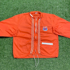 Puede incluir: Chaqueta de nylon naranja con ribetes blancos y cierre de cremallera. La chaqueta tiene un pequeño parche en el pecho izquierdo con el texto "MINNEAPOLIS" en azul.