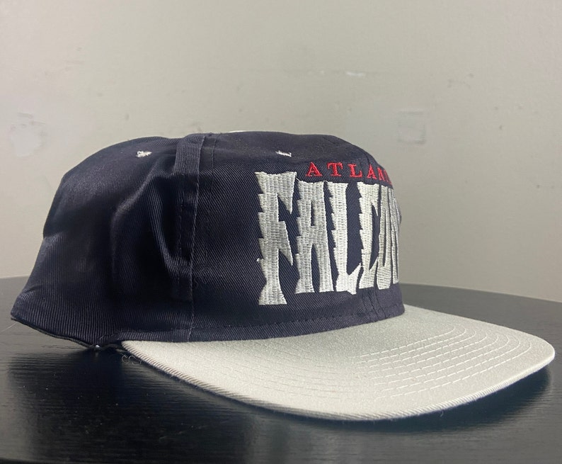May include: A black and grey baseball cap with the text "ATLANTA FALCONS" embroidered in white on the front.
