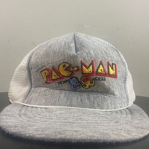 May include: A grey and white trucker hat with a Pac-Man logo embroidered on the front. The logo includes the text "PAC-MAN" and "MIDWAY 1983".