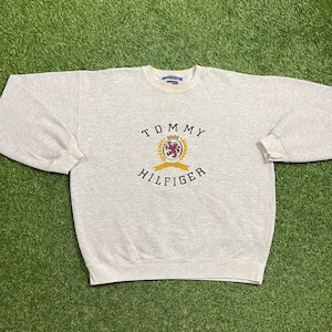 May include: A gray sweatshirt with a Tommy Hilfiger logo featuring a crest with a lion and the words "Tommy Hilfiger" in black.