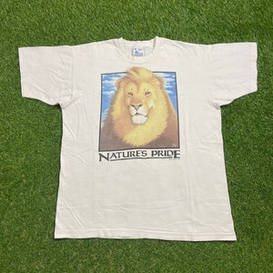 May include: White t-shirt with a graphic of a lion's head with a blue sky and clouds in the background. The text "NATURE'S PRIDE" is printed below the image.