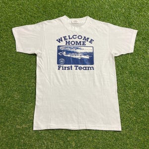 May include: White t-shirt with navy blue text and graphic. The text reads "WELCOME HOME" above a navy blue image of a ship, with "First Team" below. The shirt is made of a soft material.