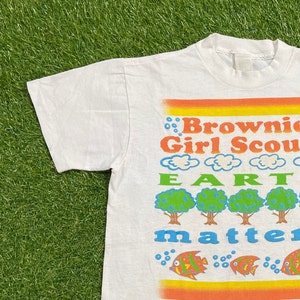 K&ouml;nnte beinhalten: Wei&szlig;es T-Shirt mit einem bunten Grafikdesign. Die Grafik zeigt den Text "Brownie Girl Scout Earth Matters" mit B&auml;umen und Fischen.