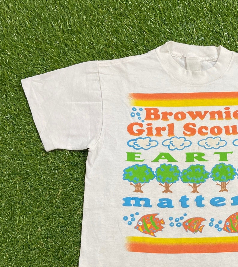 K&ouml;nnte beinhalten: Wei&szlig;es T-Shirt mit einem bunten Grafikdesign. Die Grafik zeigt den Text "Brownie Girl Scout Earth Matters" mit B&auml;umen und Fischen.