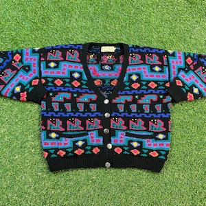 以下が含まれることがあります： カラフルな幾何学模様の黒いカーディガン。 模様には、ターコイズ、赤、青、黄色の形が含まれています。 カーディガンはVネック、半袖、シルバーのボタンが付いています。