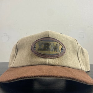 May include: A tan and brown baseball cap with a brown leather oval patch that says "IBM" in raised letters.