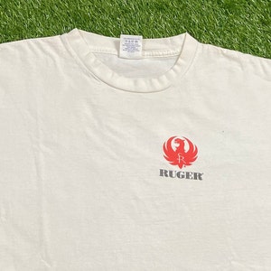 K&ouml;nnte beinhalten: Wei&szlig;es T-Shirt mit einem roten Ruger-Logo, das einen stilisierten Vogel mit einer Waffe in seinen Krallen zeigt. Der Text "RUGER" ist in Grau unter dem Logo gedruckt.