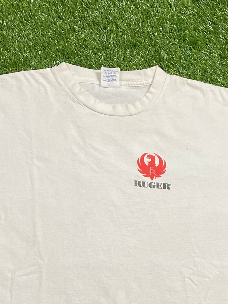 K&ouml;nnte beinhalten: Wei&szlig;es T-Shirt mit einem roten Ruger-Logo, das einen stilisierten Vogel mit einer Waffe in seinen Krallen zeigt. Der Text "RUGER" ist in Grau unter dem Logo gedruckt.