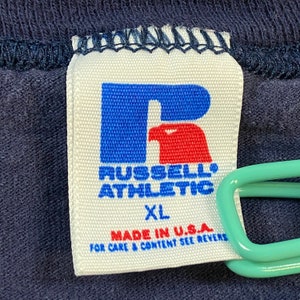May include: White tag with a blue "R" logo with a red bird inside. The text "Russell Athletic" is below the logo. The tag also says "XL" and "Made in U.S.A. For care & content see reverse."