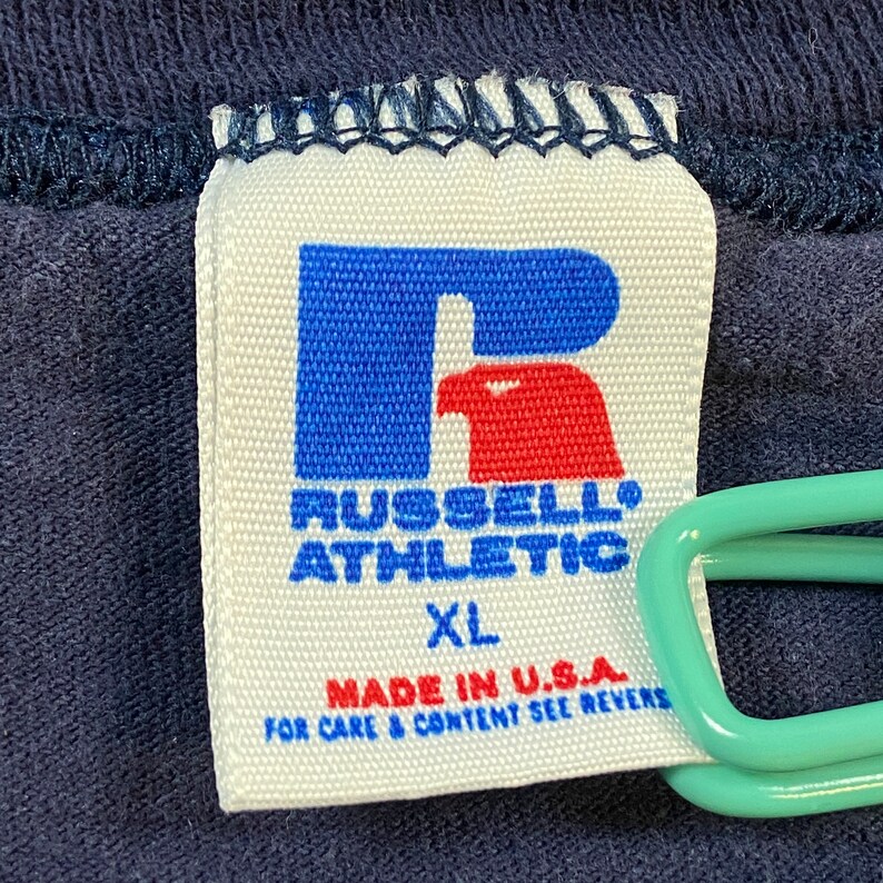 May include: White tag with a blue "R" logo with a red bird inside. The text "Russell Athletic" is below the logo. The tag also says "XL" and "Made in U.S.A. For care & content see reverse."