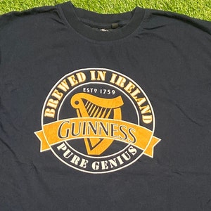 May include: A dark blue t-shirt with a yellow and black circular graphic that says "Brewed in Ireland" with a harp inside and "Guinness Pure Genius" below.