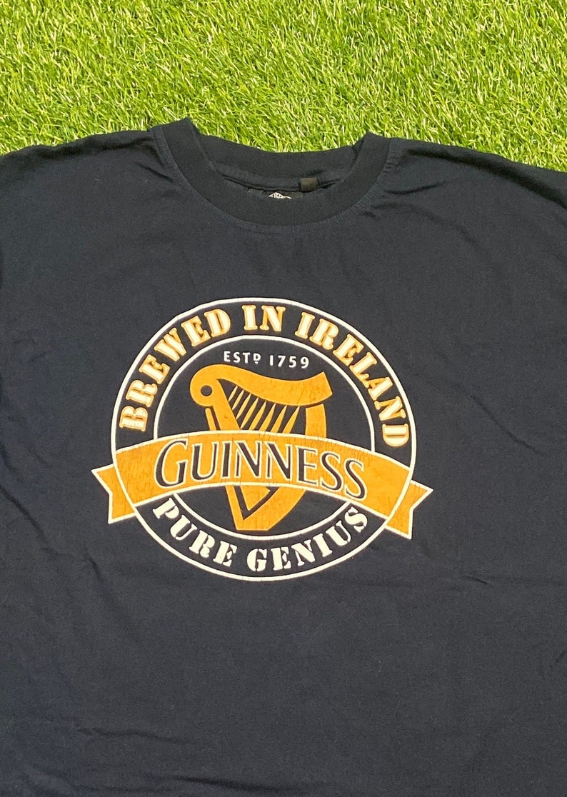 May include: A dark blue t-shirt with a yellow and black circular graphic that says "Brewed in Ireland" with a harp inside and "Guinness Pure Genius" below.