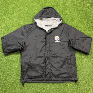 May include: A black hooded windbreaker jacket featuring a grey jersey lining and a Pittsburgh Steelers logo on the left chest. The jacket has a snap-button front closure, long sleeves with elastic cuffs, and a drawstring hem for an adjustable fit.