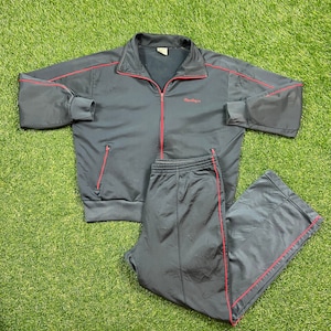 Könnte beinhalten: Ein schwarzer Trainingsanzug mit roten Paspeln. Die Jacke hat einen Reißverschluss und ein Logo mit der Aufschrift "MaxSport".