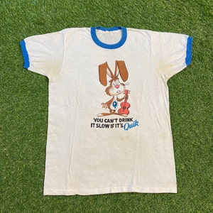 Puede incluir: Camiseta blanca con un ribete azul. La camiseta presenta un conejo de dibujos animados sosteniendo una guitarra y el texto "You can't drink it slow if it's Quik."