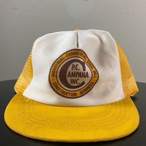 Puede incluir: Un gorro de camionero blanco y amarillo con un logotipo marrón que dice "P.C. Ampana Inc. Industrial - Commercial - Steel Mill - Construction - Services".