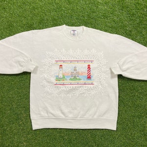 May include: White sweatshirt with a lace design featuring three lighthouses. The lighthouses are red, white, and blue.