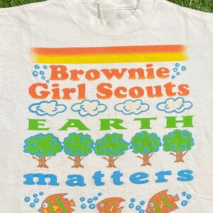 K&ouml;nnte beinhalten: Wei&szlig;es T-Shirt mit einem bunten Grafikdesign, auf dem "Brownie Girl Scouts Earth Matters" steht. Die Grafik zeigt Wolken, B&auml;ume und Fische.