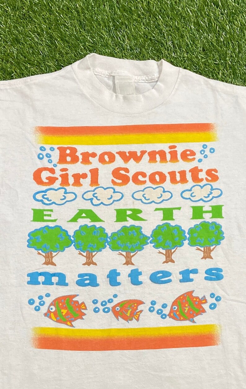 K&ouml;nnte beinhalten: Wei&szlig;es T-Shirt mit einem bunten Grafikdesign, auf dem "Brownie Girl Scouts Earth Matters" steht. Die Grafik zeigt Wolken, B&auml;ume und Fische.