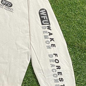 Może przedstawiać: Kremowa koszulka z długim rękawem z czarnym napisem "WFU WAKE FOREST DEMON DEACONS" na rękawie. Koszulka ma r&oacute;wnież logo "WFU" na piersi. Koszulka jest wykonana z miękkiego materiału.
