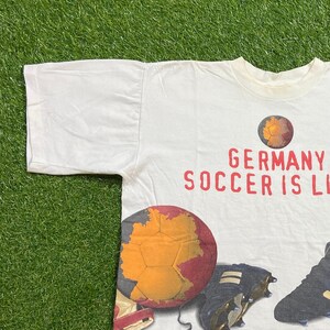 May include: White t-shirt with a graphic print of a soccer ball with a map of Germany on it. The text "GERMANY SOCCER IS LIFE" is printed in red on the shirt.