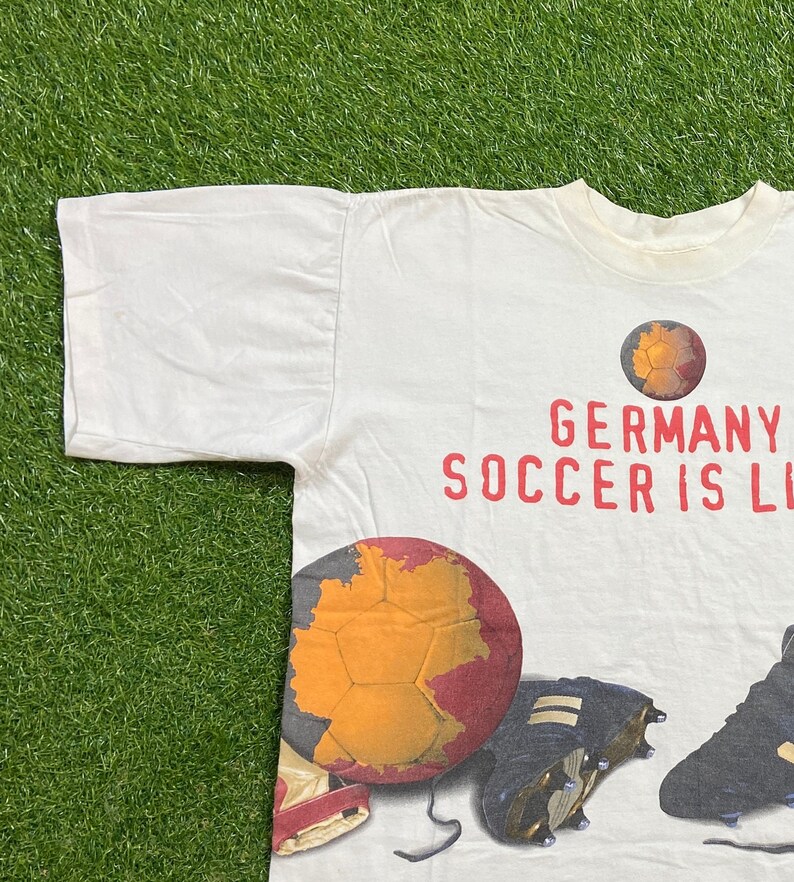 May include: White t-shirt with a graphic print of a soccer ball with a map of Germany on it. The text "GERMANY SOCCER IS LIFE" is printed in red on the shirt.