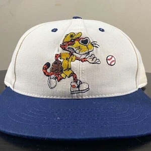 Pode incluir: Boné de beisebol branco com aba azul. A frente do boné apresenta um desenho animado bordado de um guepardo vestindo um uniforme de beisebol e segurando um taco. O guepardo corre em direção a uma bola de beisebol.