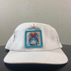 May include: White baseball cap with a blue and white embroidered patch featuring a colourful geometric design and the text "PAX/I TM".