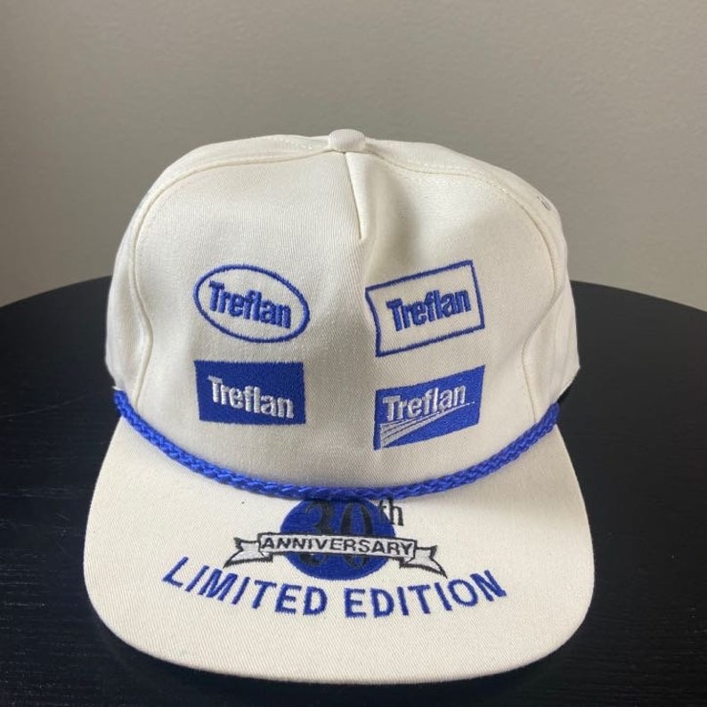 May include: White baseball cap with blue rope trim and blue embroidered logos that say "Treflan" and "20th Anniversary Limited Edition".
