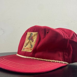 Op de afbeelding: Rode baseballpet met een witte touwrand en een gele patch met een man in een rode jas die loopt. De patch zegt "Johnnie Walker Red".