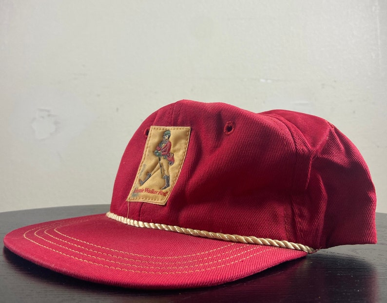 Op de afbeelding: Rode baseballpet met een witte touwrand en een gele patch met een man in een rode jas die loopt. De patch zegt "Johnnie Walker Red".