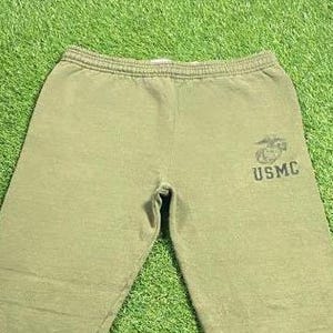 May include: Olive green sweatpants with an elastic waistband. The right leg features a USMC emblem and text in black. The pants are made of a soft, comfortable material, suitable for casual wear.