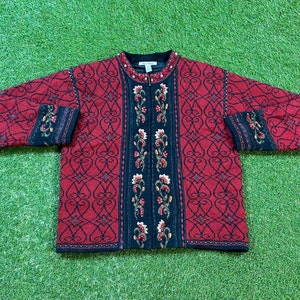 May include: A red and black patterned zip-up sweater with floral embroidery. The sweater features a black collar and cuffs, with a detailed floral design down the front and on the sleeves. The background is a green surface.