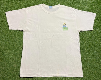 May include: A white t-shirt with a crew neck and short sleeves. The shirt features a small graphic on the front of a cartoon character wearing a hat, with the text "My Band Paradise" in a green rectangle.