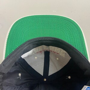 May include: The inside of a black and green baseball cap. The cap has a white brim and a white label with the text "Made in USA" on it.