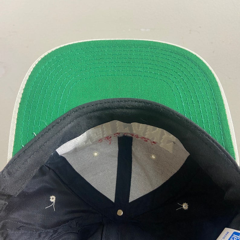 May include: The inside of a black and green baseball cap. The cap has a white brim and a white label with the text "Made in USA" on it.