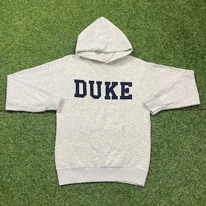 May include: A light gray long-sleeve hooded sweatshirt featuring the word DUKE printed in dark navy blue block letters across the chest. The garment includes a front kangaroo pocket, ribbed cuffs at the sleeves, and a ribbed hem at the bottom.