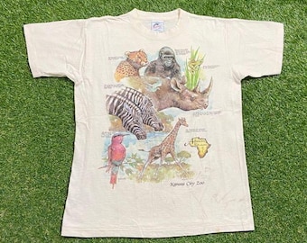 May include: Cream-colored t-shirt featuring a colorful graphic of various African animals, including a zebra, giraffe, and rhino. The design also includes a map of Africa and the text "Kansas City Zoo".