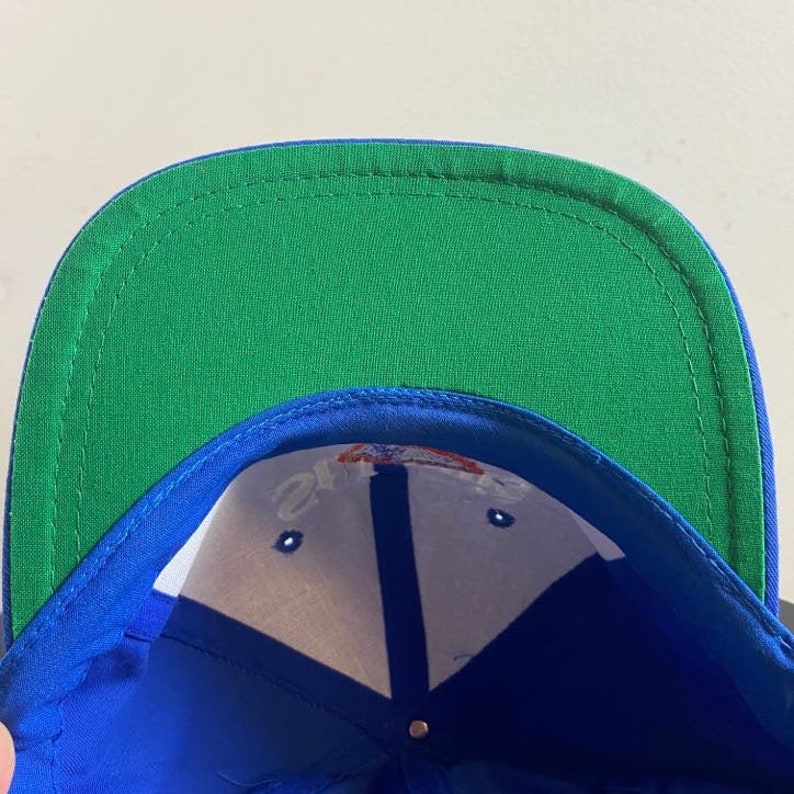 K&ouml;nnte beinhalten: Blaue und gr&uuml;ne Baseballkappe mit wei&szlig;em Innenfutter. Die Kappe hat ein wei&szlig;es Logo mit roten und blauen Akzenten.