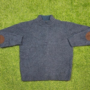 以下が含まれることがあります： モックネックとジッパーが付いた濃い青色のニットセーター。セーターの肘には茶色の革のパッチが付いています。