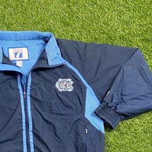 Peut inclure: Une veste coupe-vent bleu marine avec une fermeture &eacute;clair et des bordures bleu clair. La veste a un logo sur la poitrine gauche qui dit "Carolina Tar Heels".