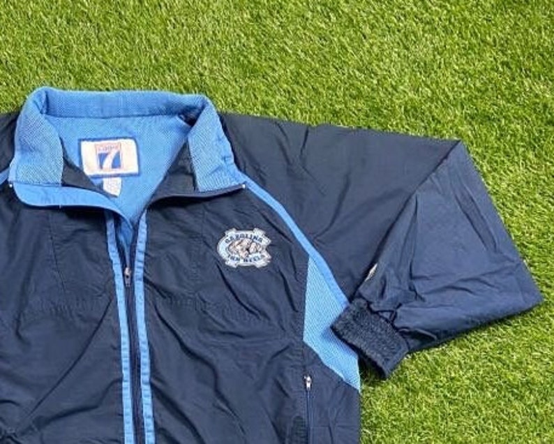 Peut inclure: Une veste coupe-vent bleu marine avec une fermeture &eacute;clair et des bordures bleu clair. La veste a un logo sur la poitrine gauche qui dit "Carolina Tar Heels".