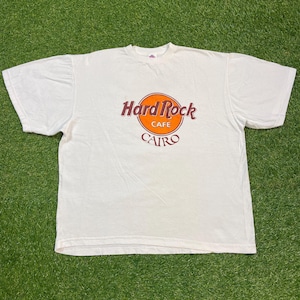 May include: White t-shirt featuring the Hard Rock Cafe logo with an orange circle and the text "Hard Rock Cafe CAIRO" in brown. The shirt is made of a soft material and has short sleeves.