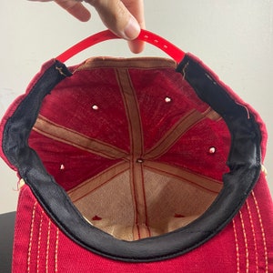 Op de afbeelding: Rode baseballpet met een zwarte klep en een beige binnenkant. De pet heeft een rood stikseldetail rond de klep.