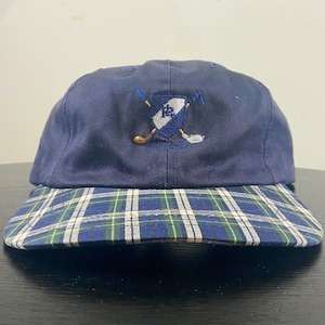 May include: A navy blue baseball cap with a plaid brim. The cap features an embroidered logo of a shield with crossed golf clubs and the letter "R" inside.